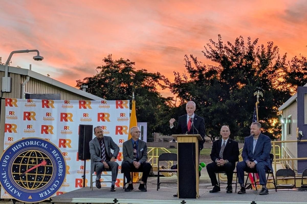 The grand opening of the National UFO Historical Records Center on the Martin Luther King Jr. Elementary School campus in Rio Rancho, New Mexico, October 2024. The center seal stands at left against a Rio Rancho Public Schools backdrop, with one of the portable classroom buildings that houses the archive at right. The Sandoval Signpost, which published the photograph, identifies the speaker at the podium as executive director David Marler.