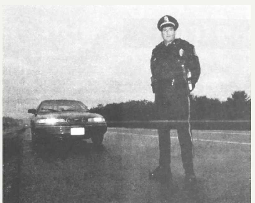 Vintage black-and-white press photograph of a uniformed police officer standing on a roadside beside his patrol car, the image used to illustrate the Kerman, California case of officer Manuel Amparano, who reported being burned by a low hovering glowing sphere on 13 May 1978. This is a real period photograph, not a CGI render or artist's impression; Amparano took no photograph of the object itself.