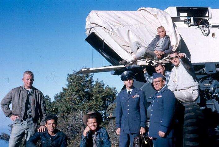 Real 1964 photograph of the Air Force tracking-telescope crew at the Big Sur site on Anderson Peak. The covered Boston University tracking telescope sits on its mount; two officers in dress blues stand front center, one carrying a briefcase, with enlisted crew and analysts around the rig on the brushy coastal mountaintop. Per the source, the group includes operations analyst Kingston George (who later argued the natural-decoy explanation) and camera officer Bob Jacobs. This is a documentary photograph of the team and equipment, not the disputed film, which was never released.