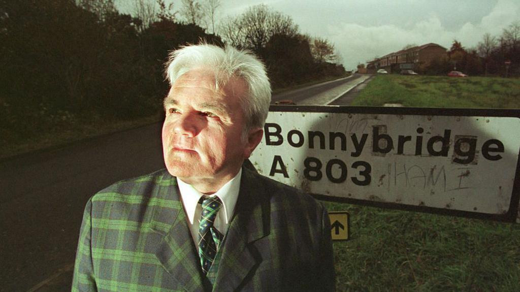 Councillor Billy Buchanan photographed beside the weathered Bonnybridge road sign on the A803. This is a press photograph of the elected member who collected and forwarded residents' reports, not a photograph of anything that was reported in the sky. No image of the Bonnybridge objects with a provenance this archive can verify exists, and the hero deliberately shows the documentary side of the case rather than a UFO picture.