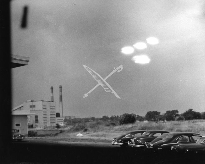 The original 16 July 1952 U.S. Coast Guard photograph taken by Seaman Shell R. Alpert from the photo lab at Coast Guard Air Station Salem, Massachusetts. Four roughly elliptical lights hang in the upper sky above the station's power-plant smokestacks and a lot of parked cars. The white "X" was added on the archival print to mark the four objects.