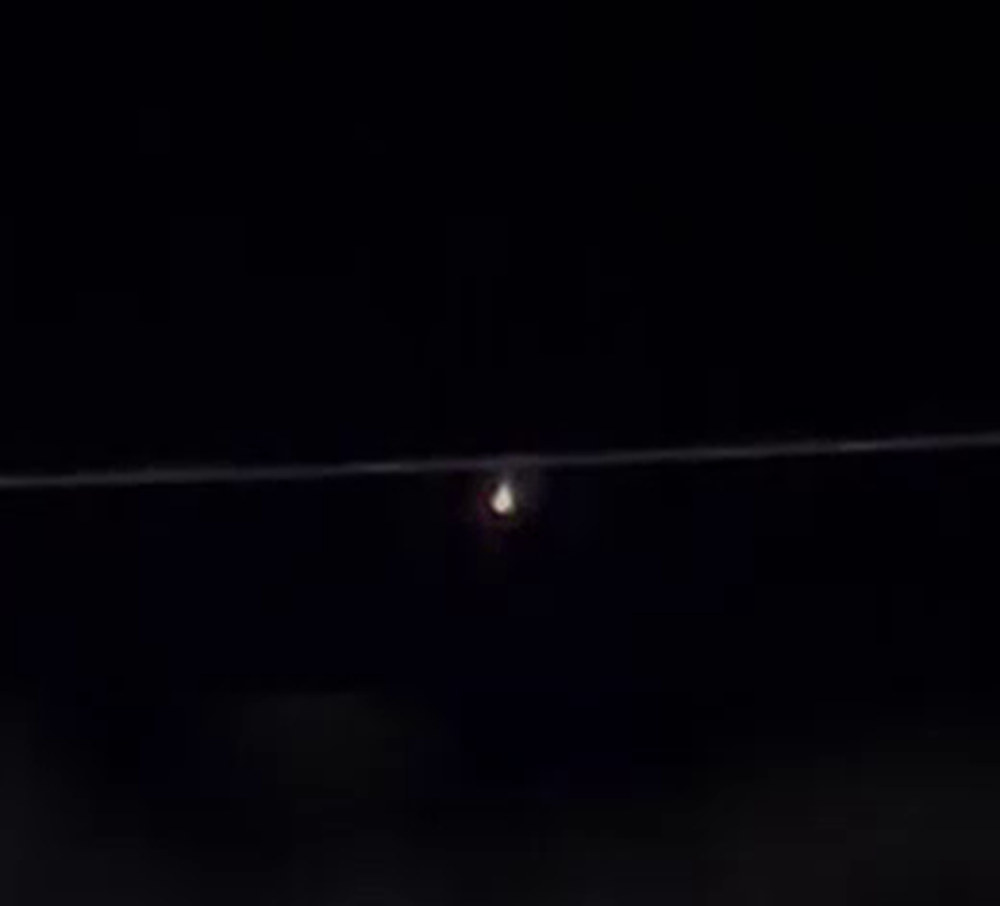 The object at normal exposure about thirty seconds into LLE-UAP-PR002, cropped and enlarged. A single power line crosses beneath it. The core is white with a warm fringe and a short downward tail, the appearance of a bright point source at high digital zoom on a phone camera.