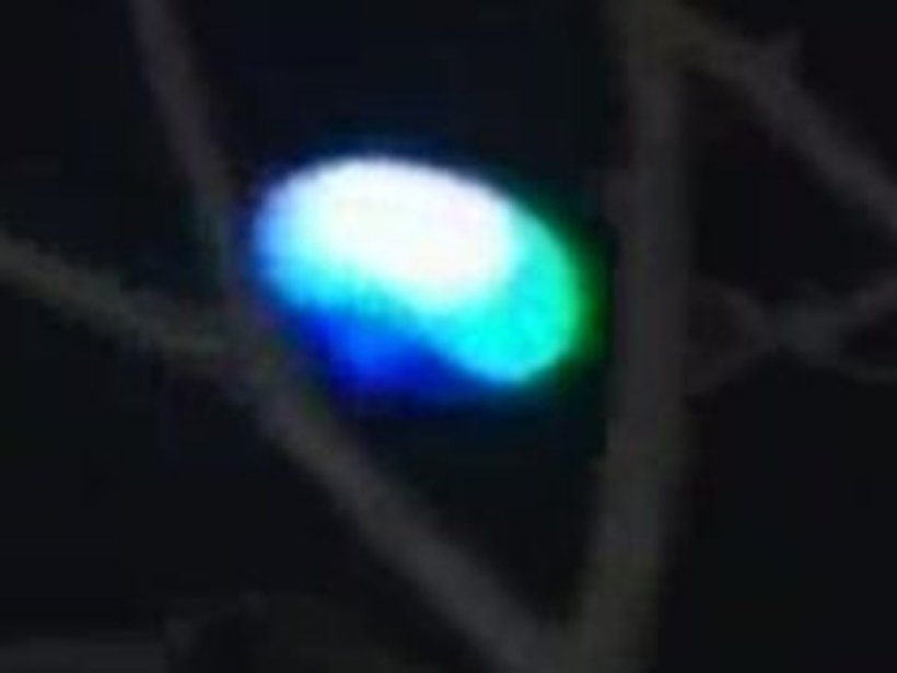 A still frame from the original witness cell-phone video, showing the glowing oval object over Winsted, Connecticut. The light glows white at the centre and is ringed with blue and green, framed by bare winter tree branches. This is a real frame from the 2011 footage, not a recreation or illustration; the strong colour fringing is partly an artifact of the low-resolution phone camera.
