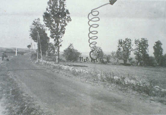 A real black and white photograph of the Cussac site taken from the children's viewpoint on the D57, showing the road, the verge and the line of hedge trees behind which the object was reported. The sphere and its helical departure path have been DRAWN onto the photograph by the investigator according to the witnesses' own indications; the spiral and the small dark shape at its top are added ink, not photographed objects. Made during Claude de Saint-Etienne's site investigation in late summer 1968.