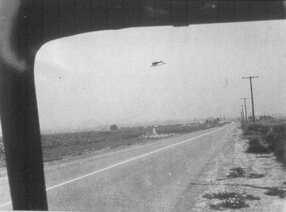 Heflin Photo 1, the first of the four frames, taken through the windshield of his Orange County Road Department van on Myford Road: the hat-shaped, dome-topped disc with its dark midsection band hovering over the road, telephone poles and the distant Santa Ana Freeway visible below.