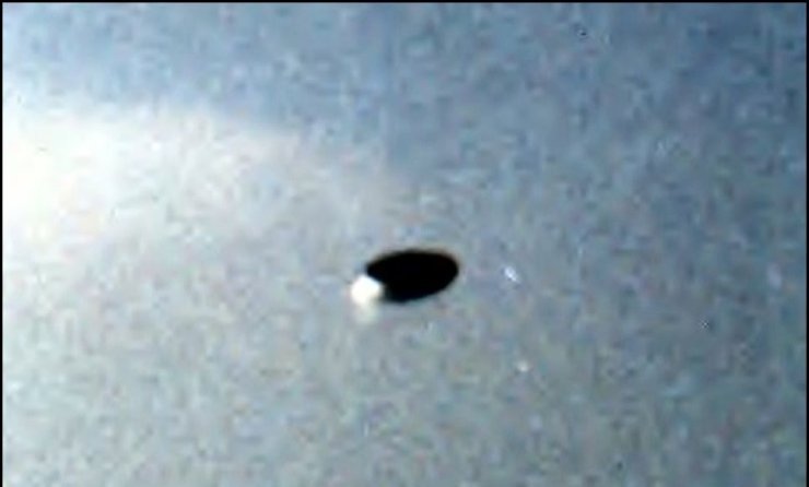 The enhanced and enlarged crop of the Érpatak photograph, showing the dark, flattened disc seen close to edge-on against a grey autumn sky, with a small bright highlight at its lower left where the sun appears to catch the surface. This is the cropped enlargement that circulates in the case literature, not the full original frame that also showed Lajos Kosina's girlfriend posing in the garden. The visible "enhanced / enlarged" line at the bottom is a UFO Casebook processing label, not part of the original negative.