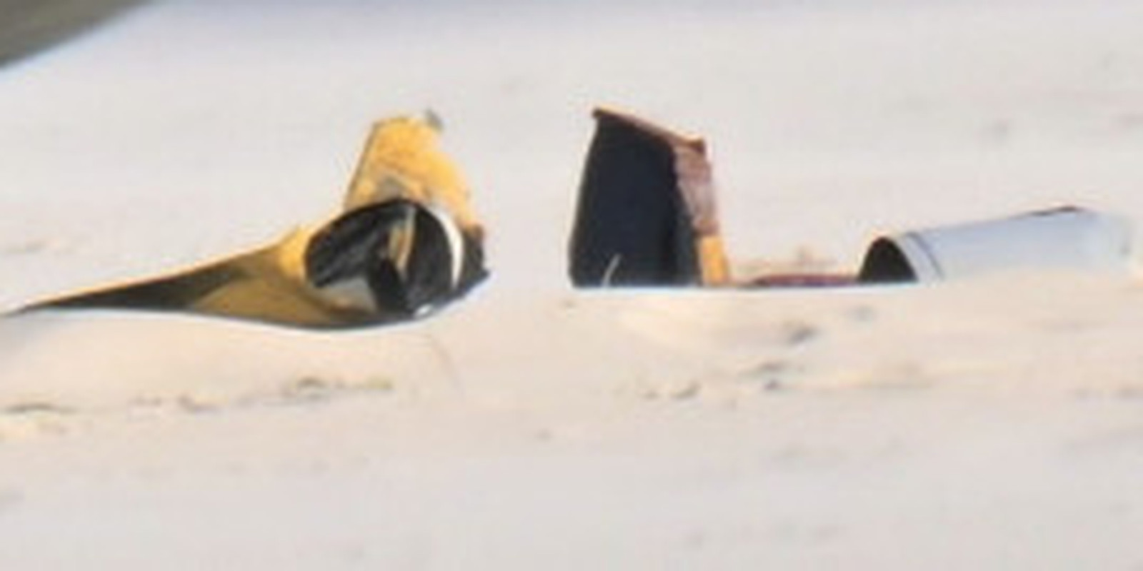 Structural fragments on the dry sand at the right of the same photograph, enlarged. A tapered yellow and tan piece with a dark hollow interior, a dark angular fragment whose break edge shows a layered sandwich construction, and a pale broken cylindrical tube. All are hollow and thin walled, which is small unmanned airframe construction. This is the material the original emergency call described as partly buried in the sand.