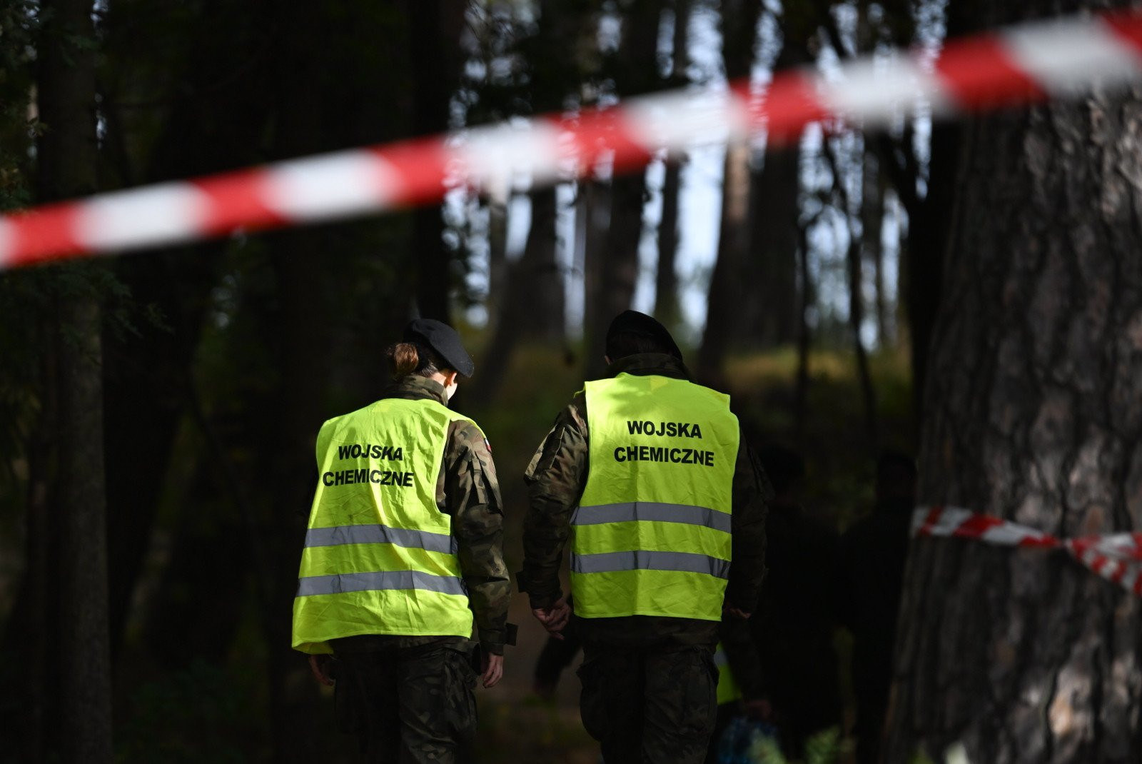 Two members of the Polish Chemical Troops, in vests reading WOJSKA CHEMICZNE, inside the red and white cordon in the pines behind the beach. This frame documents the hazard screening that the Military Police spokesman described, the pass that eliminated biological, chemical and radiological hazards and ruled out explosives before the object could be approached as ordinary debris.