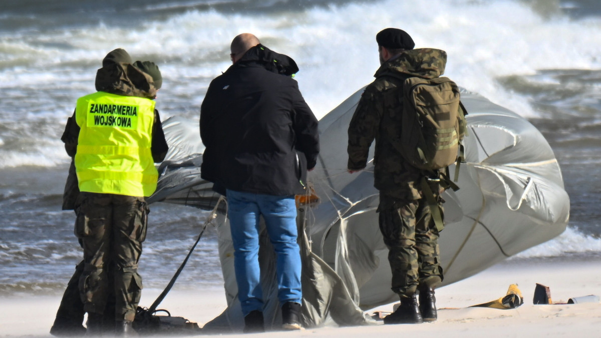 Military Police and a soldier on the sand at Karwia beside the recovered material. The large pale canopy spread across the wet sand is the clearest element in any published photograph of this case, and enlargement shows panelled fabric, shroud lines and an orange component. This is a press photograph of the recovery, not of the object in flight, and no photograph of the object before it came ashore exists.