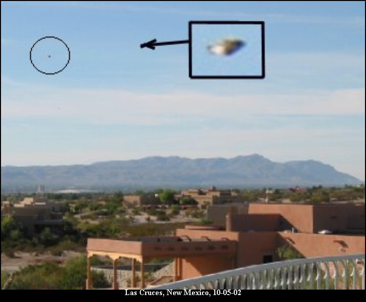 The canonical UFO Casebook composite of the 5 October 2002 Las Cruces photograph. It shows the full Canon G2 frame, a daytime landscape looking west across the Mesilla Valley toward a low desert range, with adobe rooftops in the foreground. The fainter second mark is ringed in the upper left, and a black callout box at upper centre enlarges the brighter "metallic disc" object, an arrow tying it back to its position in the sky. The callout enlargement is the website's own crop, not a separate photograph; the two objects were not seen by eye and only appeared on later computer enlargement.