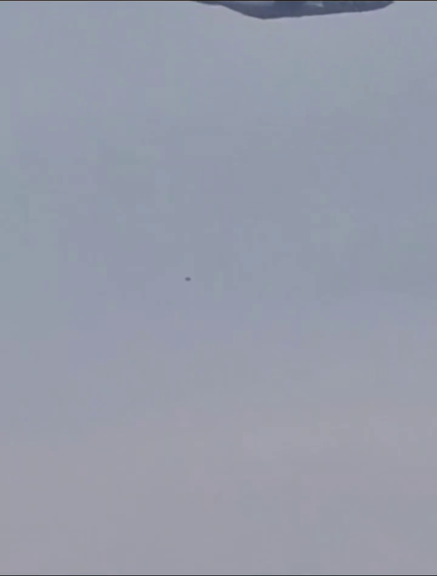 The moment that produced the headlines, shown without enhancement at twice native size. A transport aircraft crosses the top edge of the frame, its underside clipped, and the small dark object sits well below it near the centre. This is the geometry every outlet described as an object hanging beneath the aircraft. Note that the apparent pairing is a line of sight effect: nothing in a single camera view establishes that the two are at the same distance.