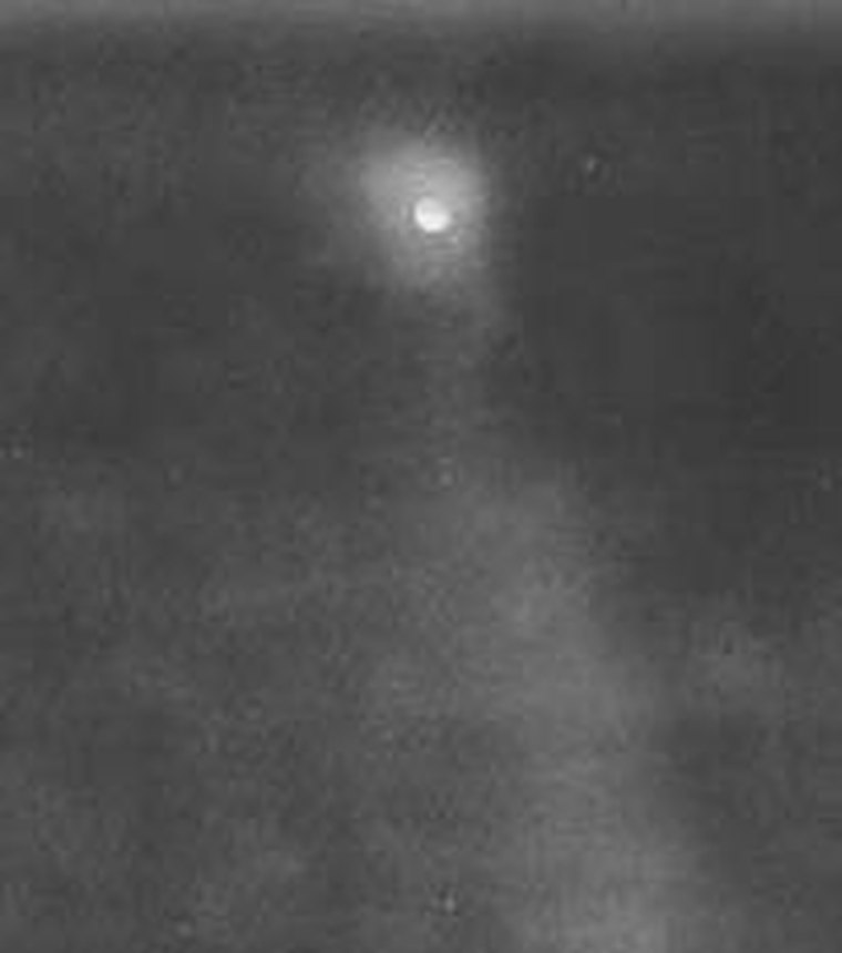 A 1949 photograph of the Norwood object: a round glowing disc at the apex of a faint searchlight beam against a grainy night sky. This is the original press photograph reproduced in Leonard Stringfield's 1957 book and later examined by Dr. Richard Haines, not a recreation or a render.