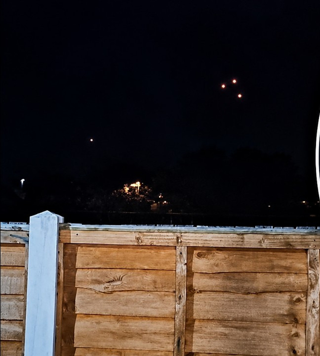 The wide frame published with the Wolverhampton footage, showing the three amber lights in their irregular triangle high in the twilight sky, a fourth isolated amber light lower and to the left, and the garden fence and treeline of the filming position. The still is taken from the aggregator upload that Coast to Coast AM embedded with the witness account.