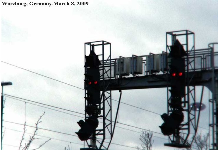 The single witness photograph, captioned "Wurzburg, Germany-March 8, 2009." The frame is dominated by a railway signal gantry over the tracks, with red rail signal lamps glowing against the dusk cloud. The alleged disk is the small faint grey shape low in the right of the frame, near the right-hand support pillar. This is the actual cell-phone image as reported, not a recreation or render.